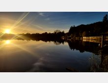 Ein ruhiger See bei Sonnenuntergang mit Spiegelung des Himmels und der umliegenden Bäume im Wasser. Am Ufer befinden sich Bäume, vereinzelte Gebäude und ein kleines Steg- oder Geländerfragment im Vordergrund rechts.