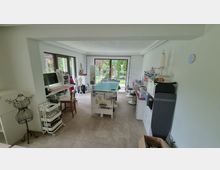 Heller Arbeitsraum mit großen Fensterfronten, die auf einen Garten blicken, ausgestattet mit einem Schneidertisch, mehreren Regalen, Nähmaschinen und Stoffrollen. Ein Schaufensterpuppe und verschiedene Werkzeuge für Handarbeit sind verteilt, darunter ein Bügelbrett an der Wand.