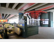 U-Bahn-Station mit grünen Kacheln und einer Rolltreppe im Vordergrund, darüber eine Betondecke mit roten Elementen. Im Hintergrund ein kreisförmiger Eingang mit roten und schwarzen Designelementen, beschriftet mit „Bierpinsel“.