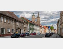 Straßenszene einer Kleinstadt mit Reihen von Mehrfamilienhäusern in verschiedenen Farben und einer Kirche mit zwei Türmen im Hintergrund. Am Straßenrand parken mehrere Autos, und Laternenpfähle säumen die Gehwege.
