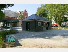 Kleines einstöckiges Gebäude mit dunkler Backsteinfassade, Fenstern und einem Glasbereich, umgeben von einem gepflasterten Platz mit Bänken und Bäumen. Im Hintergrund sind eine erhöhte Bahntrasse mit vorbeifahrendem Zug und ein rotes Backsteingebäude zu sehen.