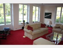 Wohnzimmer mit roten Teppichboden, cremefarbenen Sofas, roten Zierkissen und einem Glastisch in der Mitte. Große Fenster lassen viel Tageslicht herein und bieten einen Blick auf einen grünen Garten.