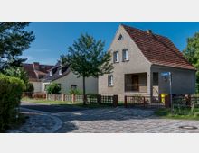 Einfamilienhaus mit rotem Satteldach und hellem Putz, eingebettet in eine Wohnsiedlung mit gepflasterten Gehwegen und Vorgärten. Im Vordergrund ein Straßenschild mit der Aufschrift „Saartandstr.“ und eine gelbe Mülltonne am Eingang.