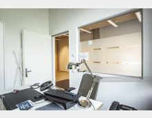 Büroarbeitsplatz mit Schreibtisch, auf dem sich eine Metall-Schreibtischlampe, ein Telefon, eine Tastatur und Büroartikel befinden. Im Hintergrund sind eine offene Tür, eine Garderobe und ein Fenster mit Sichtschutzstreifen zu sehen, durch das ein angrenzender Raum erkennbar ist.