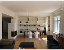 Wohnzimmer mit hellen Wänden, einem großen Bücherregal an der Rückwand und einem Holzboden. Ein rechteckiger Holztisch steht vor dem Regal, umgeben von zwei weißen Stühlen, mit modernen Möbeln wie einer schwarzen Couch, einem Sessel und einem kleinen Kamin an der linken Wand.