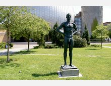 Bronzestatue einer stehenden männlichen Figur auf einem Sockel, umgeben von einer grünen Parklandschaft mit Rasen, Bäumen und Büschen; im Hintergrund ein modernes Gebäude mit einer großen, silbernen Kuppel und weiteren Gebäudeteilen.