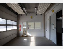 Industriehalle mit einem großen Rolltor auf der linken Seite, einer weißen Tür und einem Fenster zu einem angrenzenden Raum in der Mitte. An der Wand hängen rote Kästen und Hinweisbeschilderungen, ergänzt durch eine graue Tür am rechten Bildrand.