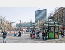 Straßenszene in einer belebten Stadt mit einem modernen mehrstöckigen Gebäude im Hintergrund, auf dessen Dach das Mercedes-Benz-Logo zu sehen ist. Im Vordergrund steht ein kleiner Pavillon mit der Aufschrift „Blumen“, umgeben von Passanten, Fahrrädern und Straßenlaternen; Schilder und Werbetafeln prägen das Stadtbild.