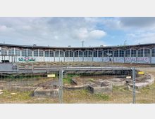 Rundbau eines historischen Lokschuppens mit großen, bogenförmigen Fenstern und zugemauerten Toren, teilweise mit Graffiti bedeckt. Im Vordergrund eine mit einem Metallzaun abgegrenzte Drehscheibe für Schienenfahrzeuge, umgeben von überwachsenem Gelände.