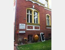 Rotes Ziegelgebäude mit gelben Fensterrahmen und leicht geschwungenen Bögen über den Fenstern. Im Vordergrund stehen Dekorationsgegenstände wie ein großer Keramiktopf, eine Figur und ein Verkehrslicht, neben Schildern mit der Aufschrift „Richardplatz“ und „Militärischer Sicherheitsbereich!“