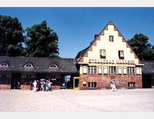Backsteinbau des Eingangsgebäudes des Strandbads Wannsee mit gelb gestrichenen Akzenten und Satteldach, flankiert von großen Bäumen im Hintergrund; vor dem Eingang befindet sich eine Gruppe von Menschen.