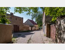 Schmale gepflasterte Gasse in einem Dorf mit alten Ziegelmauern und Gebäuden, darunter ein gelbes Haus mit rotem Dach. Im Hintergrund stehen Bäume, und Sonnenlicht fällt durch die Blätter.