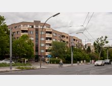 Mehrstöckiges Wohngebäude mit cremefarbenen Fassaden und Balkonen, umgeben von Bäumen und Grünflächen. Im Vordergrund eine Straße mit Straßenbahnschienen, einem Radfahrer und einigen Autos sowie einem Gehweg mit Verkehrsschildern.