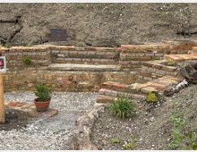 Ziegelsteinterrassen in einem Gartenbereich, eingebaut in einen leicht ansteigenden Hang. Im Vordergrund sind Kiesboden, kleinere Pflanzen sowie ein Blumentopf sichtbar, während dekorative Elemente wie Holzstücke und Metallplatten die Szenerie ergänzen.
