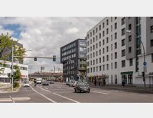 Straßenszene mit moderner Architektur, flankiert von mehrstöckigen Büro- und Wohngebäuden mit großen Fensterfronten. Im Hintergrund fährt ein ICE-Zug über eine erhöhte Schienenbrücke, während Autos und Radfahrer die Straße beleben; Fußgängerwege, Ampeln und ein Buswartehäuschen sind ebenfalls sichtbar.