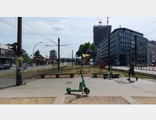 Straßenszene in einer Stadt mit Straßenbahnschienen, modernen Bürogebäuden und Kunstinstallationen in der Mitte eines kleinen Platzes. Zwei grüne E-Scooter stehen auf dem Gehweg, während eine Person vorbeigeht und Autos sowie Geschäfte im Hintergrund zu sehen sind.