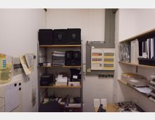 Kleiner, funktionaler Abstellraum mit Regalen voller Akten, Dokumente und Aufbewahrungsboxen. An der Wand befindet sich ein Stromverteilerkasten, daneben Notizen, Papierstapel und ein Tisch mit Kabeln und technischen Geräten.