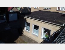 Ein flacher Anbau mit beigem Putz und drei Fenstern, die durch einfache weiße Gardinen teilweise verdeckt sind. Rechts davon fällt der Schatten eines Geländers, und im Hintergrund sind weitere Gebäude mit roten und grünen Dächern sichtbar.