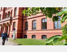 Rotes Backsteingebäude mit Rundbogenfenstern und verzierten Säulen am Eingangsbereich, umgeben von gepflegtem Grün und vereinzelten Bäumen. Im Vordergrund läuft ein Mann auf einem Gehweg vorbei, begleitet von einem Verkehrsschild und einer Informationstafel.