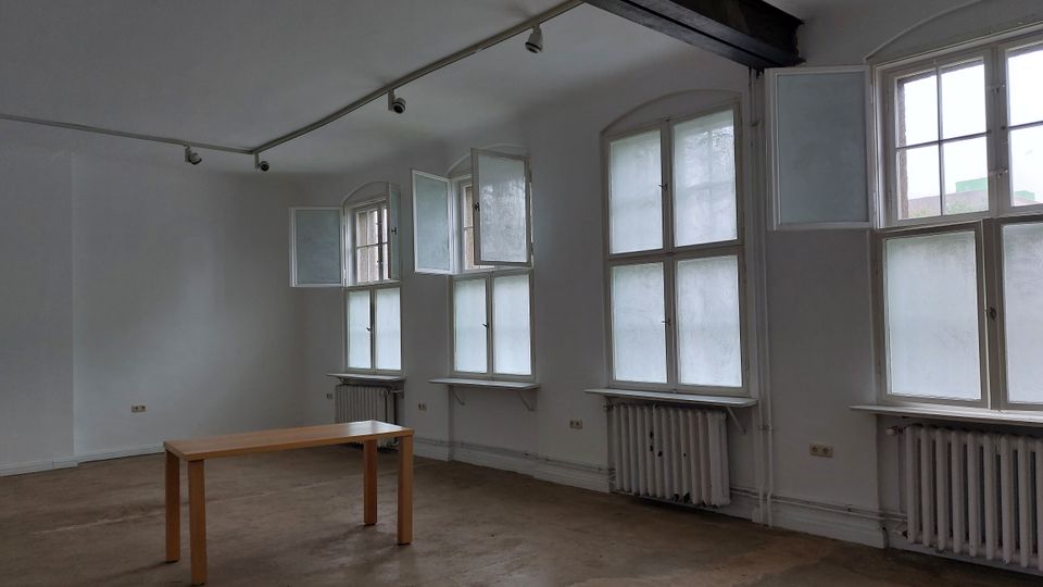 Ein schlichtes, leerstehendes Zimmer mit weiß gestrichenen Wänden, großen, hohen Fenstern mit Milchglas und geöffneten Flügeln sowie Heizkörpern unter den Fenstern. Im Raum steht ein einfacher, schmaler Holztisch auf einem Betonboden.