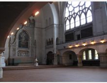 Innenansicht einer historischen Kirche mit hohen, gotischen Fenstern auf der rechten Seite, die viel Tageslicht einlassen. Links ist ein reich verziertes Altarbild in einer Wandnische zu sehen, umgeben von kunstvoller Architektur und Statuen; der Boden besteht aus großen, roten Fliesen.