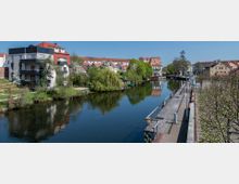 Wohnanlage und Promenade entlang eines Flusses in einer Kleinstadt. Auf der linken Seite befinden sich moderne und traditionelle Gebäude, umgeben von Bäumen, während rechts ein gepflasterter Weg mit Geländern und Sitzbänken parallel zum Wasser verläuft.