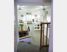 Ein modernes Büro mit weißen Wänden, offenen Regalen und mehreren Arbeitsplätzen, das von natürlichem Licht durch ein Fenster am Ende des Raumes beleuchtet wird. Im Vordergrund befindet sich ein Holztisch mit Stühlen sowie ein Treppenaufgang mit einem Holzgeländer.