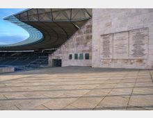 Großes Stadion mit einer geschwungenen, modernen Überdachung aus Glas und Stahl. Im Vordergrund eine breite Steinfläche vor einer hohen Natursteinfassade mit eingearbeiteten Gedenktafeln und Reliefs.