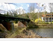 Grüne Stahlbrücke mit ornamentiertem Geländer, die über einen Fluss führt, getragen von Backsteinpfeilern; im Hintergrund sind Bäume und ein Gebäudekomplex mit weißen Fassaden und roten Dächern zu sehen.