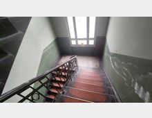 Treppenhaus in einem älteren Gebäude mit grün gestrichenen Wänden, einem schwarzen Metallgeländer und roten Stufen. Am Ende der Treppe befindet sich ein großes Fenster, das Tageslicht hereinlässt, und ein gefliestes Muster auf dem Boden.