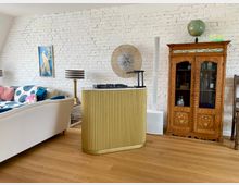 Wohnzimmer mit heller Einrichtung auf Holzboden. Eine weiße Backsteinwand dient als Hintergrund, darauf ein Bild mit einem Pfauenmotiv. Im Vordergrund befindet sich ein goldener DJ-Tisch, rechts ein hölzerner, verzierter Schrank mit Glastüren und Globus darauf, links ein helles Sofa mit bunten Kissen.