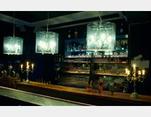 Elegant gestaltete Bar mit hölzerner Theke, großen schimmernden Kronleuchtern unter durchsichtigen Lampenschirmen und mehreren Kerzenleuchtern. Im Hintergrund gut bestückte Regale mit Gläsern und Flaschen vor einem beleuchteten Spiegel.