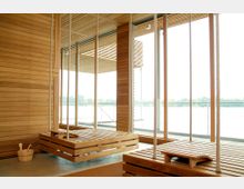 Innenansicht eines modernen Ruheraums aus Holz mit großen bodentiefen Fenstern, die einen Blick auf eine Außenterrasse und Wasserlandschaft bieten. Zwei hölzerne Schaukeln mit Seilen hängen von der Decke, daneben steht ein kleiner Holzeimer mit einer Kelle.