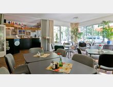 Heller Caféraum mit großen Fenstern, mehreren Tischen und Stühlen, dekoriert mit kleinen Blumenvasen und Tischdecken. Im Hintergrund befindet sich eine Theke mit Holzverkleidung und Dekoration aus internationalen Flaggen.