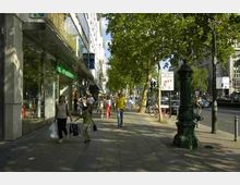 Straßenszene in einer Einkaufsstraße mit Gehweg, auf dem Fußgänger spazieren und Einkaufstaschen tragen. Links moderne Schaufensterfassaden, rechts eine Baumreihe mit einem historischen grünen Trinkbrunnen im Vordergrund.