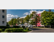 Mehrstöckige Wohngebäude mit weißen und roten Fassaden, gesäumt von Bäumen und Sträuchern, stehen entlang einer Straße mit parkenden Autos und einem Gehweg. Der Himmel ist leicht bewölkt, und die Umgebung wirkt gepflegt.