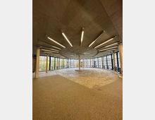 Großer, moderner Innenraum mit roh belassener Decke, schlanken Betonstützen und umlaufender Glasfassade, die einen Ausblick auf Bäume und Gebäude bietet. Die Decke ist mit langen, symmetrisch angeordneten Leuchtbalken ausgestattet, und der Boden besteht aus unterschiedlich strukturierten Oberflächen.