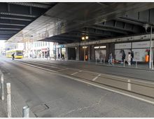 Straßenansicht unter einer Brücke mit sichtbaren Stahlträgern und Beleuchtung. Rechts befinden sich Schaufenster eines Gerry-Weber-Geschäfts, davor mehrere Personen und ein Obdachlosenlager; links fährt eine gelbe Straßenbahn auf den Gleisen.
