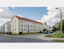 Mehrstöckiges Wohnhaus mit heller Fassade und rotem Satteldach, gelegen an einer Straßenecke mit Kopfsteinpflaster. Im Umfeld befinden sich eine kleine Rasenfläche, parkende Autos, Straßenlaternen und Bäume.