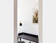 Minimalistischer Arbeitsbereich mit einem schwarzen Schreibtisch, einer gold-schwarzen Tischlampe und einem Stuhl mit grünem Stoffbezug. An der weißen Wand hängt ein Poster mit einer botanischen Illustration von Palmen.