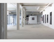 Großer, moderner Ausstellungsraum mit Betonpfeilern, glatten Böden und weißen Wänden, an denen mehrere abstrakte Kunstwerke hängen; im Raum befinden sich Skulpturen und Sitzgelegenheiten aus Metallgittern.