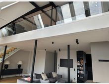 Moderner, offener Wohnraum mit hoher Decke, sichtbaren schwarzen Stahlträgern und einer Galerie mit Glasgeländer. Unten sind ein großes graues Sofa, eine Treppe zur Galerie, dezente Einbaubeleuchtung sowie ein Regalbereich neben einem Durchgang zu sehen.