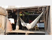 Holzstruktur eines offenen Schuppens oder Bootshauses mit Blick auf das Innere, das eine Hängematte, einen Holztisch mit Tischdecke und Stühle zeigt. Im Inneren hängen verschiedene Gegenstände und Dekorationen von der Decke, darunter Girlanden und Haushaltswerkzeuge.