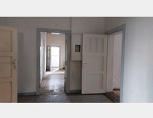 Ein leerer Innenraum mit mehreren geöffneten weißen Holztüren, die in weitere Räume führen. Der Boden besteht aus abgenutzten Fliesen, die Wände sind hell gestrichen, und das Licht fällt von einem Fenster in einem der hinteren Räume ein.