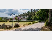 Eine ländliche Szene mit einem unbefestigten Weg, der an einer Wiese mit Steinzaun und Bäumen vorbeiführt; im Hintergrund steht ein Haus mit roten Dachziegeln und angrenzenden Nebengebäuden, umgeben von Wald. Am Weg befindet sich ein Straßenschild mit der Aufschrift „Rheinfeldene Allee“.