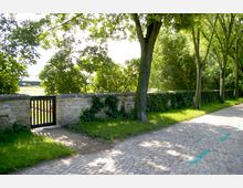 Gepflasterter Weg neben einer niedrigen, mit Efeu bewachsenen Sandsteinmauer mit Holztor; im Hintergrund Bäume und Sträucher, die teils Schatten auf den Weg werfen.