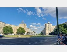 Eine breite Straßenszene mit großen, symmetrisch angeordneten Wohngebäuden im sowjetischen Baustil, flankiert von Bäumen. In der Mitte des Platzes befinden sich eine Grünfläche und ein Springbrunnen, während eine Person auf einem Fahrrad im Vordergrund zu sehen ist.
