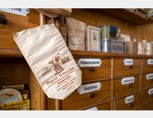 Nahaufnahme eines alten Holzregals in einem Kolonialwarenladen mit kleinen Schubladen, die Etiketten wie "Stollwerck" und "Gibzucker" tragen. Im Vordergrund hängt eine bedruckte Kaffeetüte mit der Aufschrift "Horst Fritzsche Kaffee" und einer Illustration.