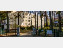 Mehrstöckiges Wohngebäude, vermutlich ein Seniorenheim, mit hellem Putz, umgeben von hohen Bäumen und einer Grünanlage. Im Vordergrund ein Eingangstor mit Hinweisschildern und ein Weg, der zum Gebäude führt.