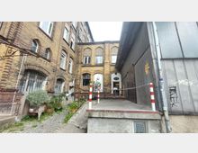 Hinterhof mit einem historischen Backsteingebäude auf der linken Seite und einer modernen Stahlfassade rechts; der zentrale Eingangsbereich ist über eine Rampe erreichbar und mit Pflanzenkübeln umgeben. Kopfsteinpflaster und eine rot-weiße Absperrung prägen den Vordergrund.